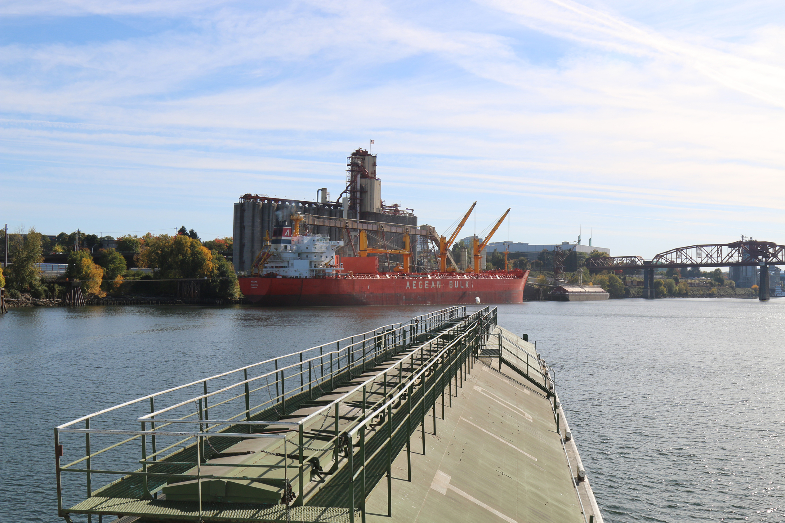 Photo taken from a tugboat pushing a grain barge down the Willamette River toward an export elevator with a bulk vessel docked and ready to be loaded.
