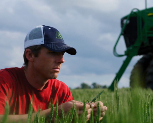 Photo of Kansas farmer Justin Knopf in his wheat field examining plant leaves for disease pressure with the front of a self-propelled application machine in the background.