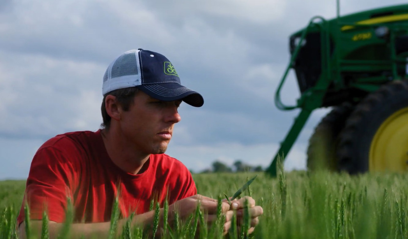 Photo of Kansas farmer Justin Knopf in his wheat field examining plant leaves for disease pressure with the front of a self-propelled application machine in the background.
