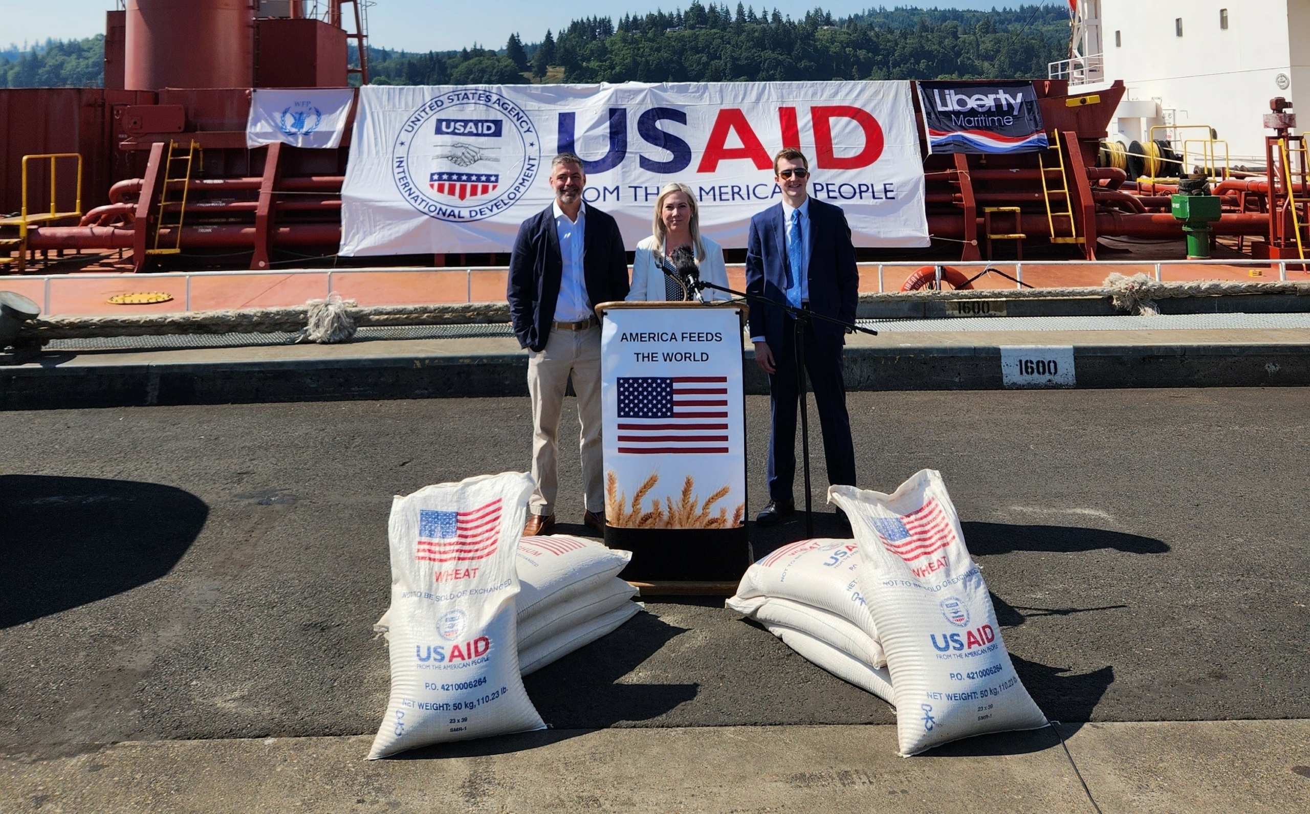 Three people stand at a podium on the dock of a grain export elevator with a sign saying USAID on a ship behind them and bags of wheat marked "USAID: Donated by the American People".